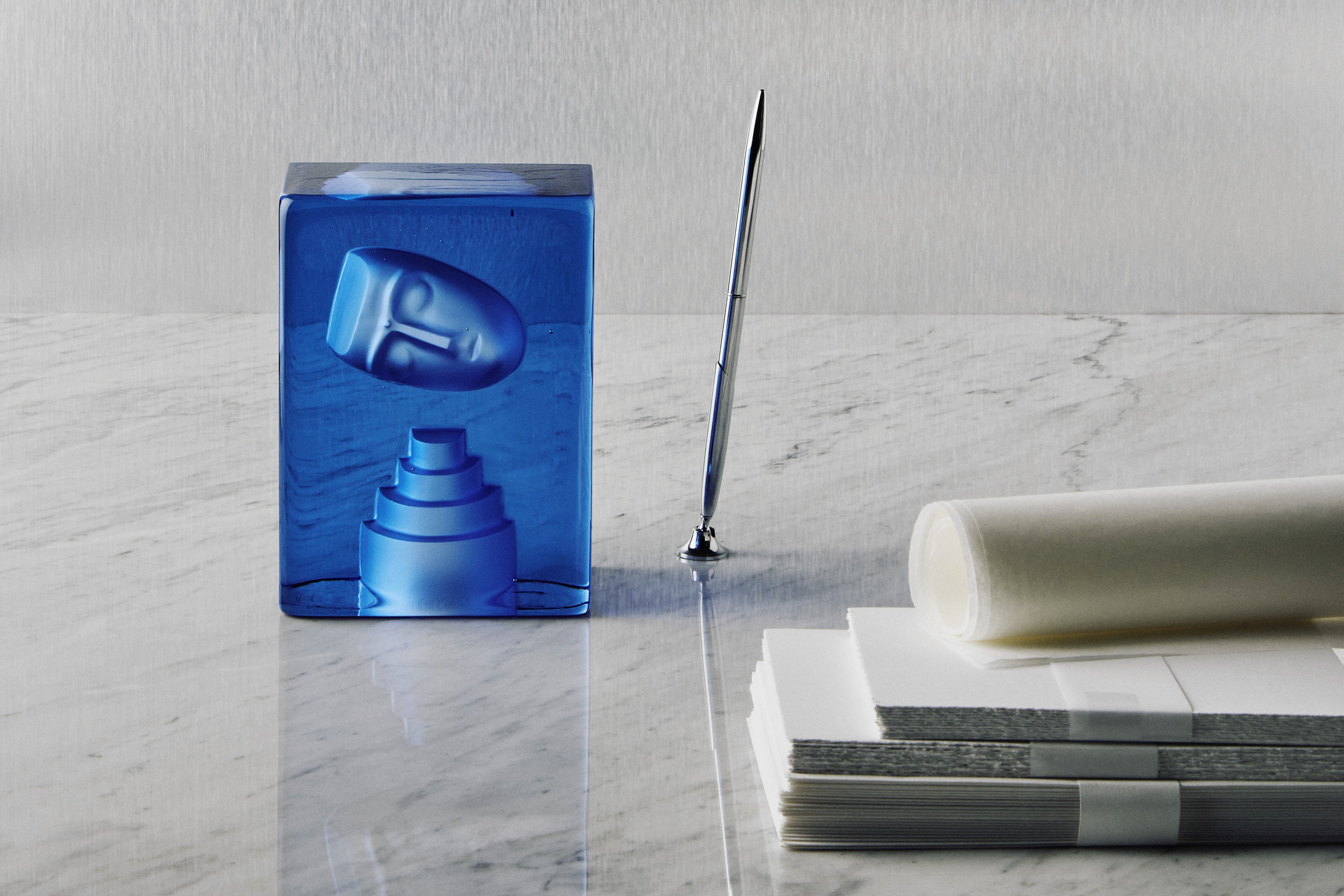 Kosta Boda Azur Blue glass sculpture on a marble surface with a pen and paper.