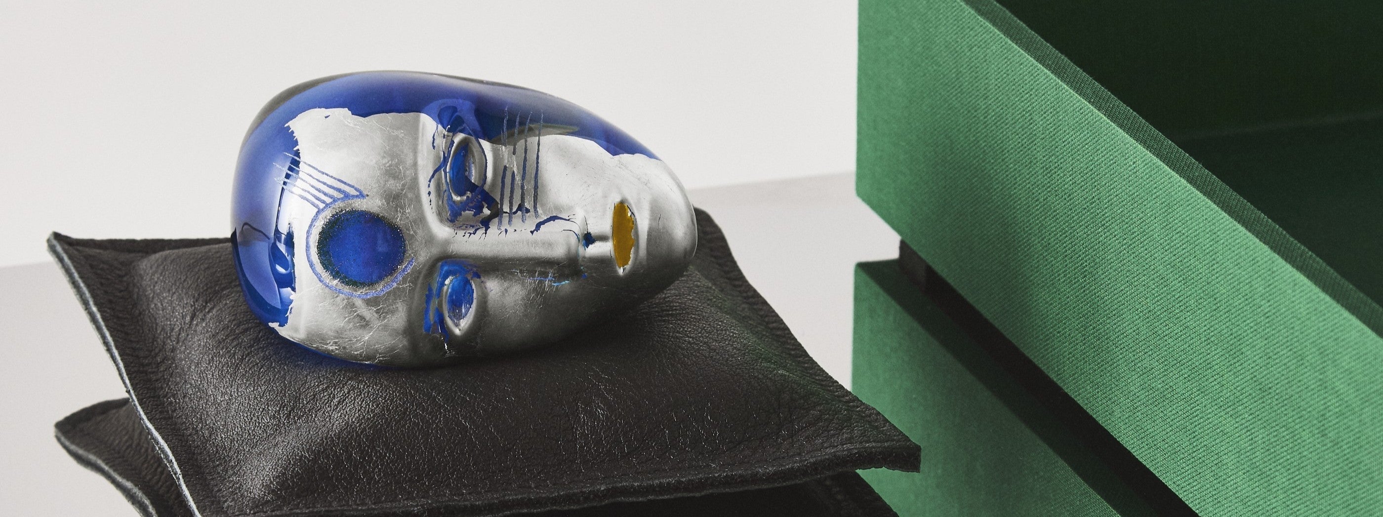 Glass head by Bertil Vallien with blue and silver colours on a black cushion against a green and white background