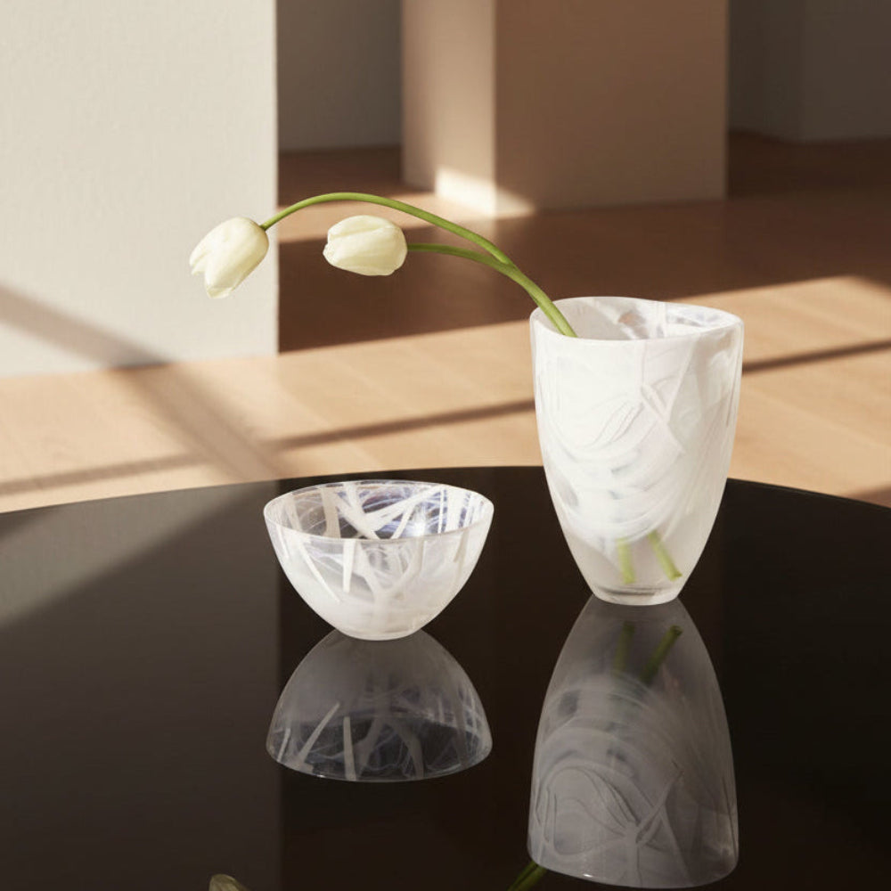 Kosta Boda Contrast white glass vases with textured, swirling patterns sit on a glossy black table. One vase is larger and holds a single white tulip, while the other is a smaller matching bowl-shaped vase.