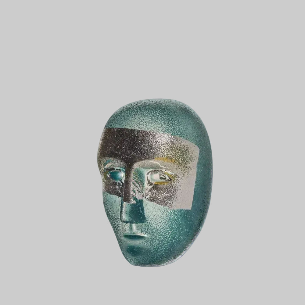 A sculptural decorative mask with a textured, metallic teal finish, featuring a black band across the forehead and a light gray square overlay on the eye area, set against a plain white background.