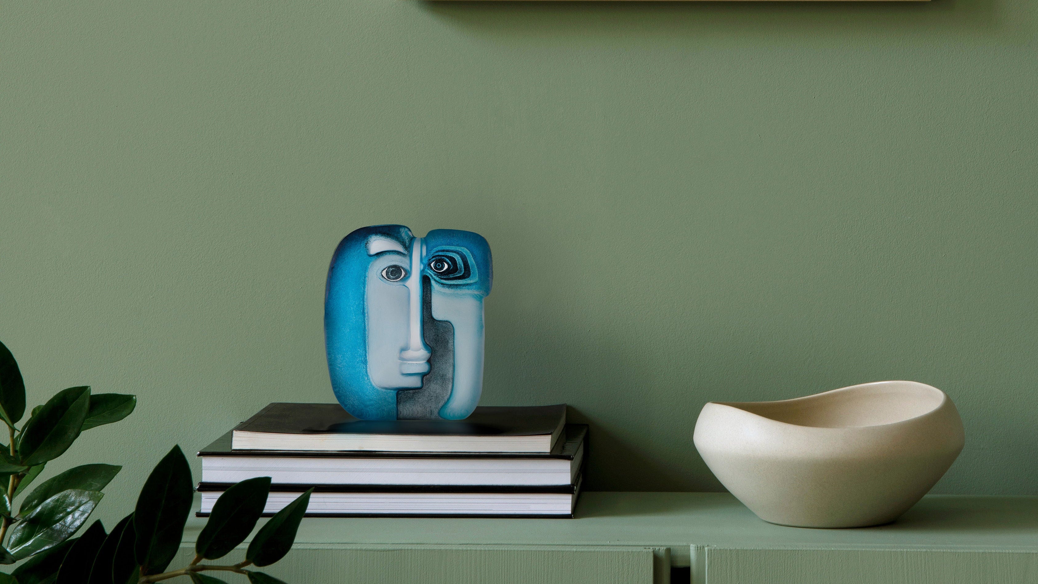 Maleras glass sculpture on books with a bowl on a green shelf against a green wall