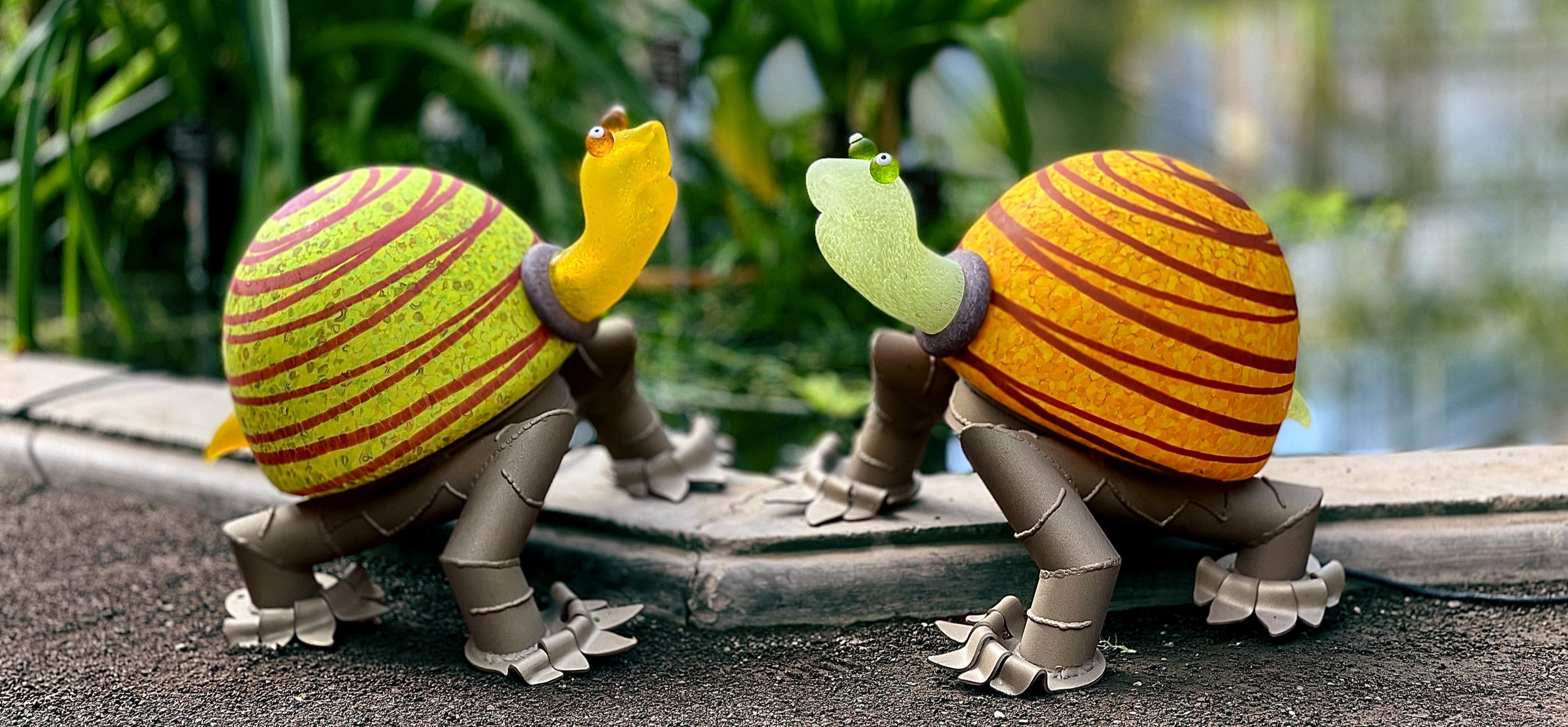 Two colorful turtle figurines on a path with greenery and water in the background