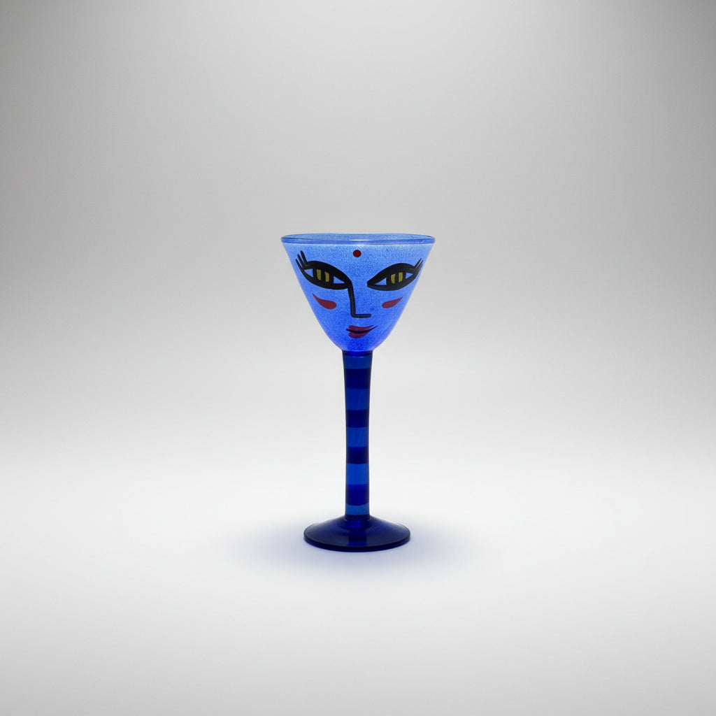 Open minds blue goblet-shaped glass candle holder with a stylized face design, standing on a white shelf against a neutral gray background.