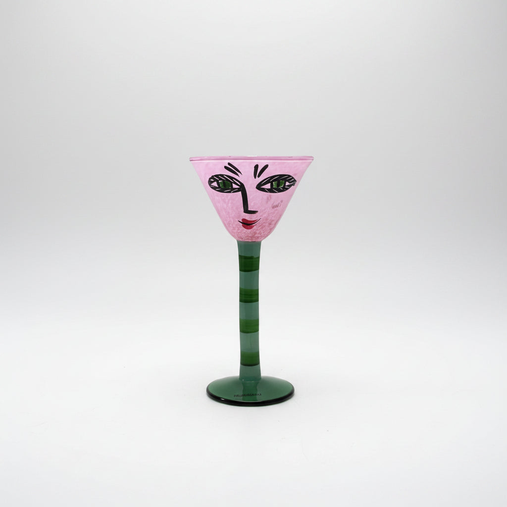 Kosta Boda Open Minds decorative pink and green glass goblet with a stylized face painted on the bowl, set against a plain white background.