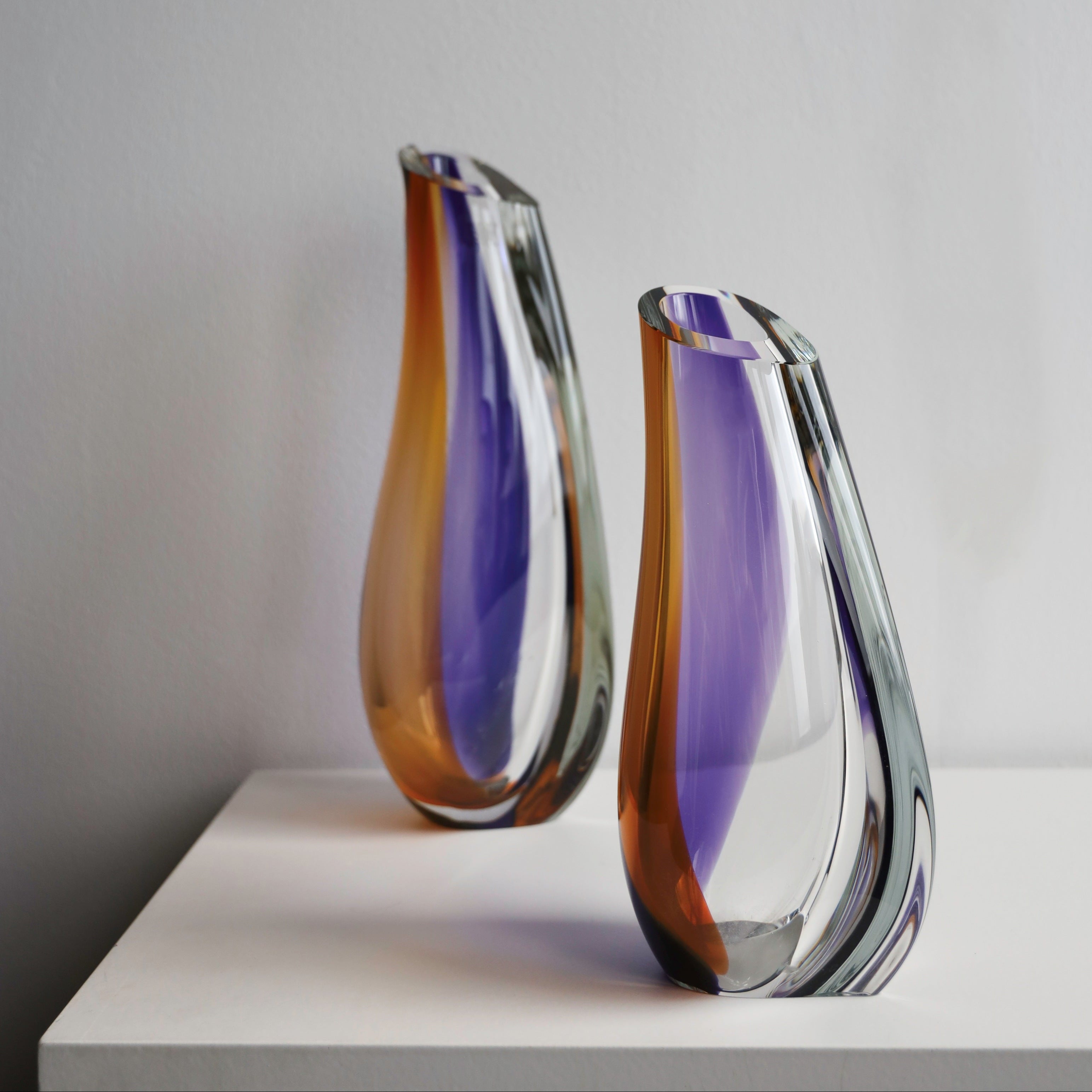 Two Kosta Boda glass vases with a gradient colour pattern on a white surface and grey background