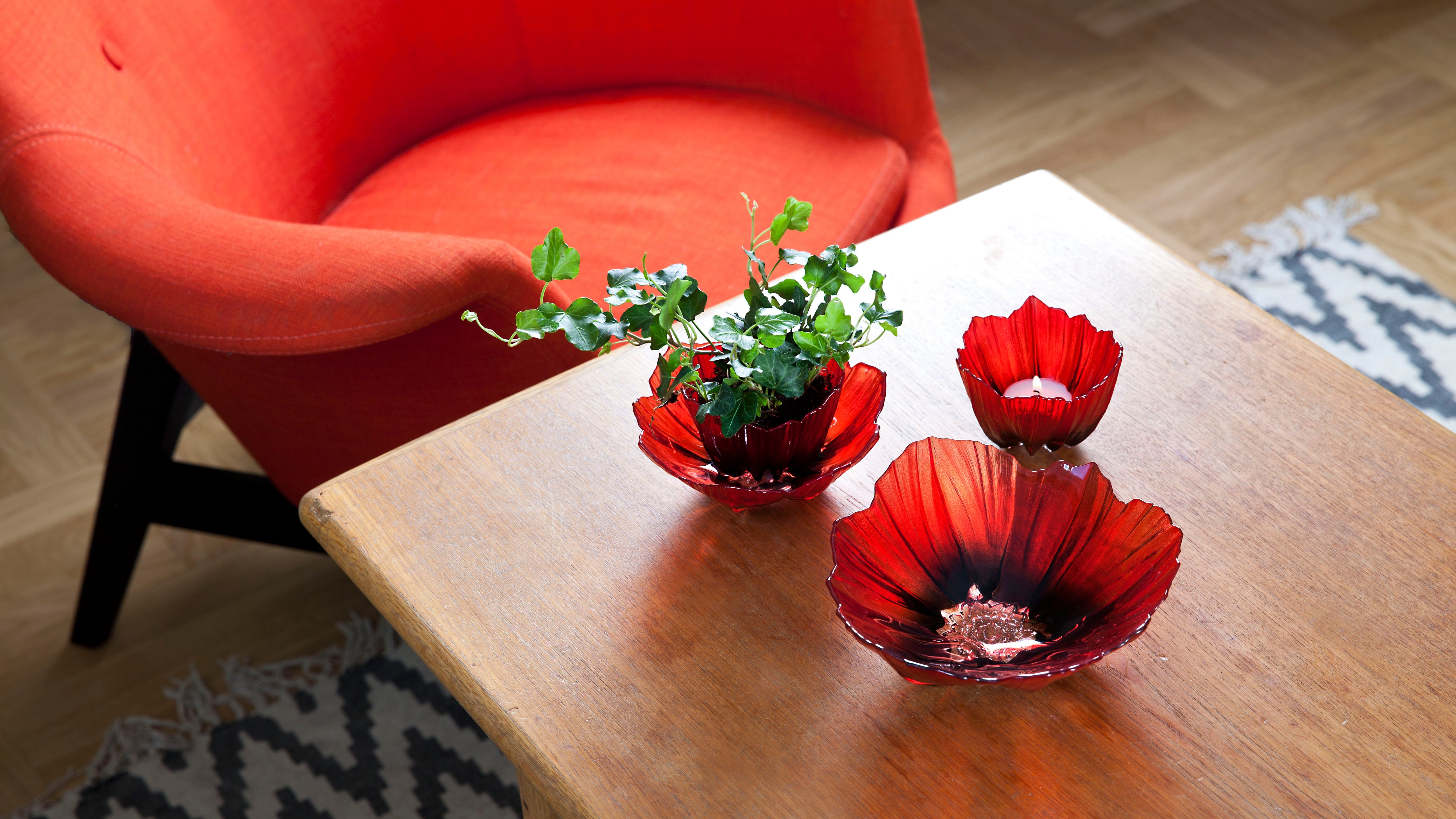 Three red glass bowls with greenery on a wooden table next to a red chair.