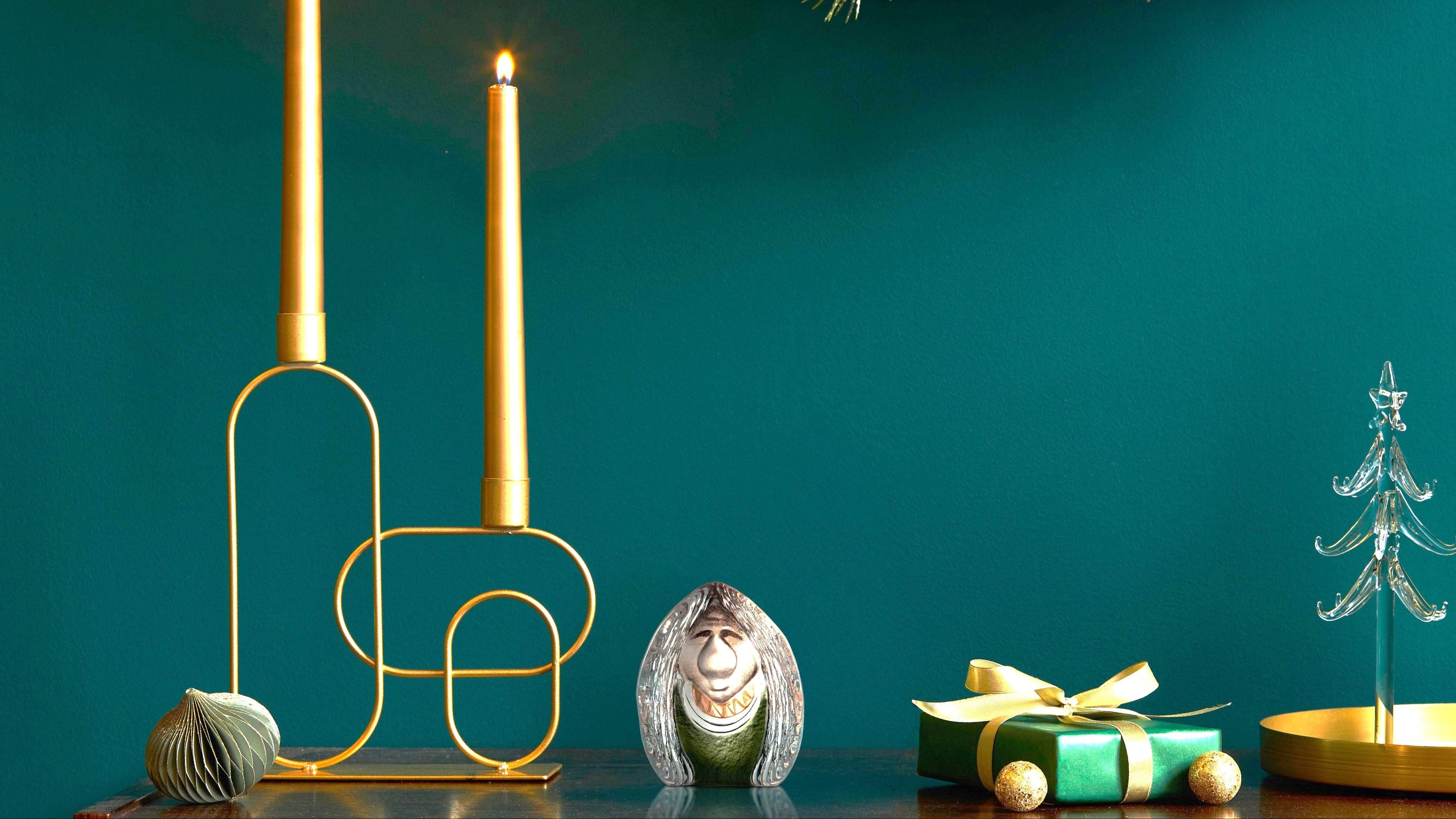 Decorative setup with gold candle holders, a small statue, wrapped gift, and Christmas tree ornament against a teal background.