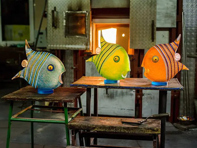 Three colorful fish-shaped lamps on wooden tables in a workshop setting.