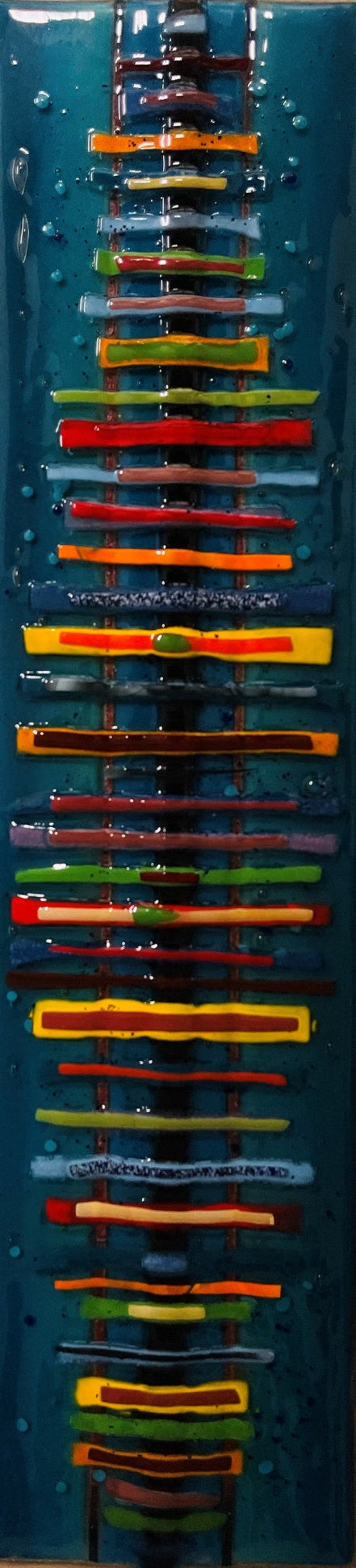 A vertical rectangular glass artwork featuring a dark teal-blue background with multiple horizontal bands of colorful glass in red, orange, yellow, green, and purple.