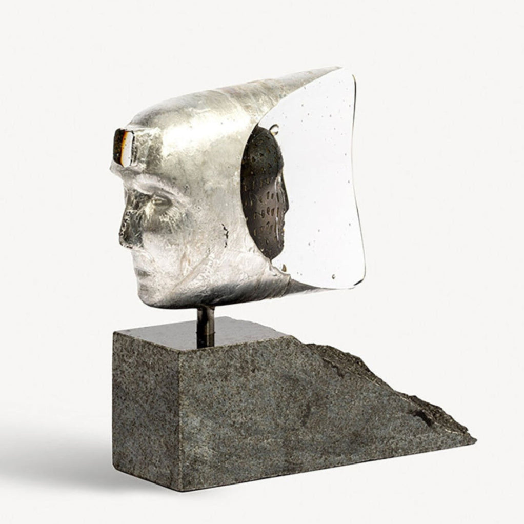 Sculpture of a head with a metallic helmet on a grey base against a white background