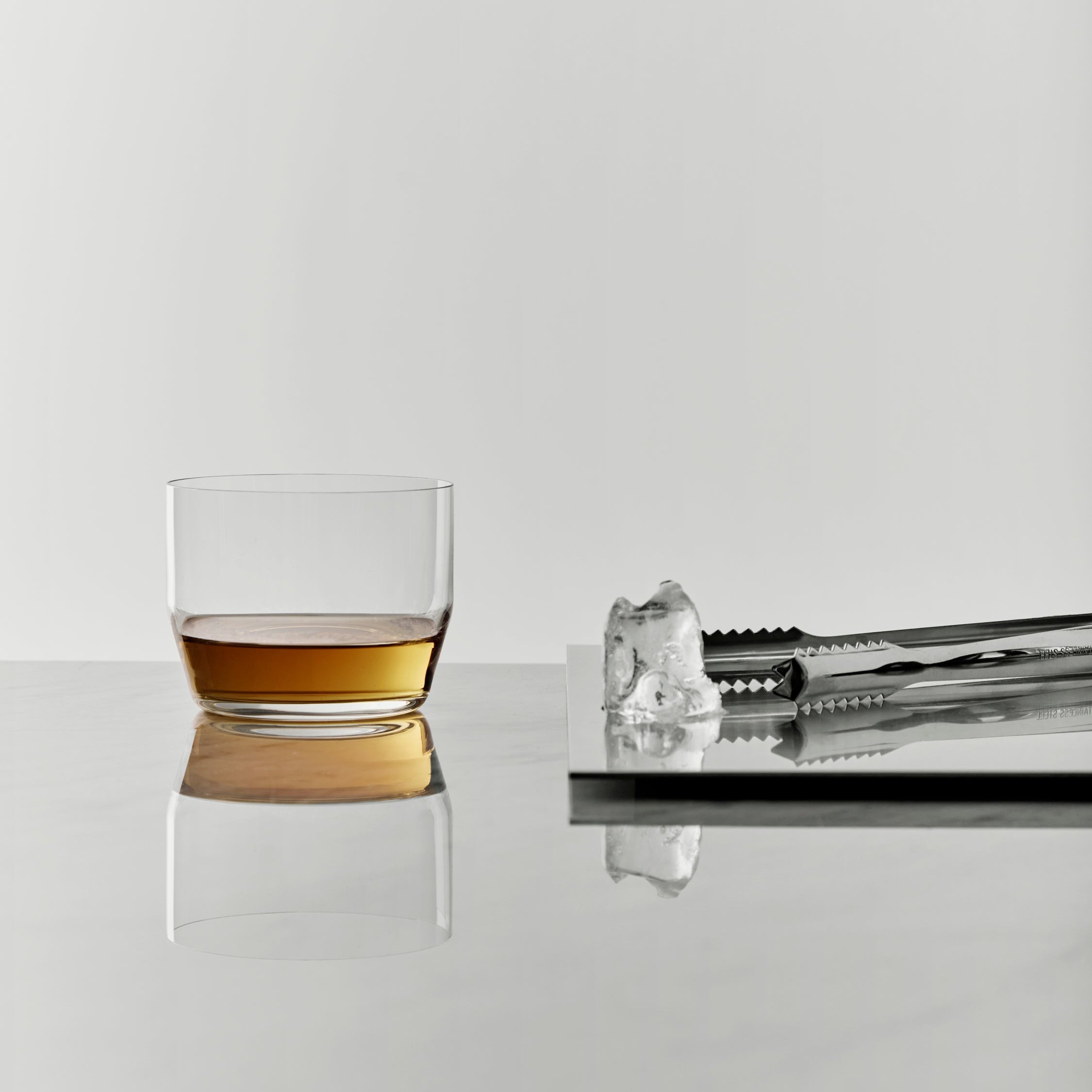 Glass of whiskey with a ice cube and tongs on a reflective surface