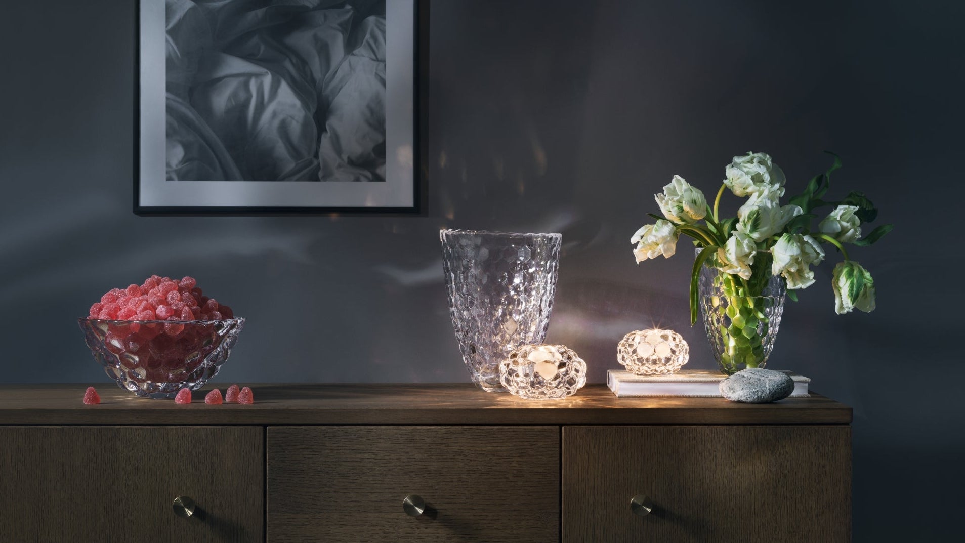 Dark wooden dresser with decorative items including a vase, flowers, and small lights against a dark wall.